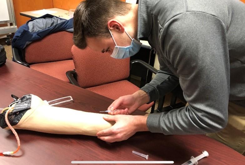 =A student uses a latex IV arm to practice administering IVs. 