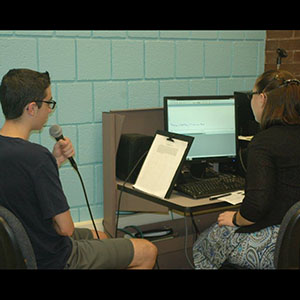 Person speaking into a microphone at a computer with a grad student reviewing results of experiment.