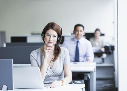 Woman in office at desk wearing headset with coworkers at desks in the background