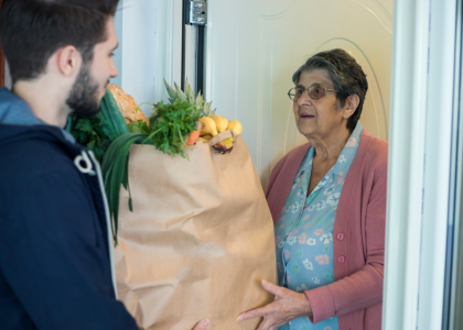 A person hands another person groceries in a paper bag at their front door. 