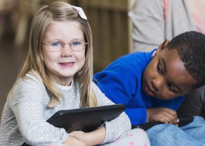 Two preschool students sit together. A White girl holds an iPad while a Black boy looks at her. 