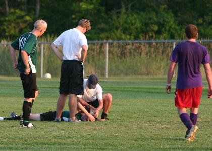 Injured soccer player laying on his back on the pitch. Concerned coaches attend to him as two players look on, concerned