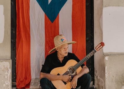 Elderly man with guitar sitting in front of Puerto Rican flag.