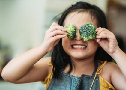 young girl holding broccoli in front of her eyes and smiling