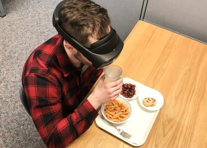 Man sits in cubicle wearing a virtual-reality headset with a meal in front of him. He is drinking from a glass.