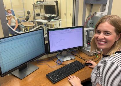 Olivia Leach smiles behind a bank of computer screens while Rachel Cottle sits behind a glass window