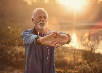 An elderly man stretches his hands before him, he is standing in front of a lake at sunset