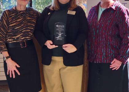 Cynthia Bittner, holding award, poses with Lisa Davis and Karen Burke in front of large glass windows.