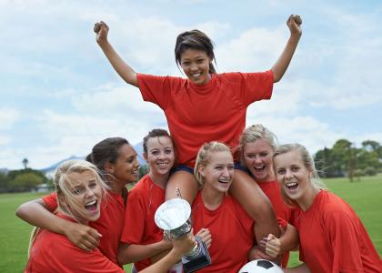 Women's sports team in a huddle, holding a trophy and raising a teammate on their shoulders.