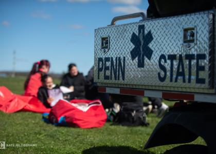 A Penn State emergency vehicle is parked on a grassy field with students sitting on the ground in the background.