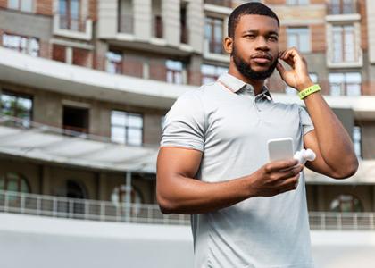 A man wearing a polo shirt and a fitness tracker on his wrist holds a smartphone while adjusting his headphones. 