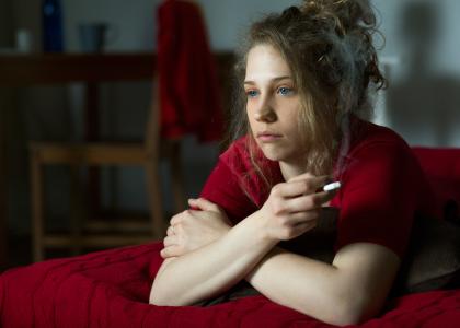 Woman laying on bed with lit cigarette looking morose