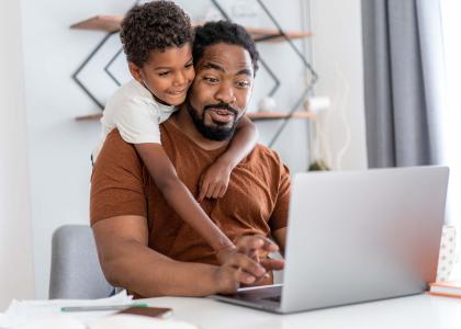 A father working at home on a laptop with his son's arms wrapped around him.