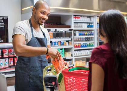 Black, college-aged man bags groceries and smiles at a customer