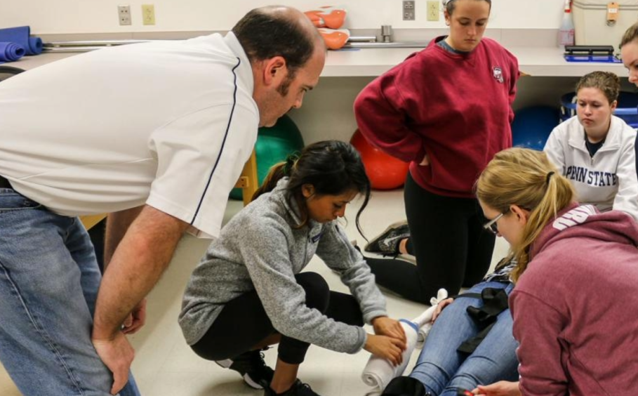 A group of students practice securing a patient to a stretcher with an instructor overlooking the process. 