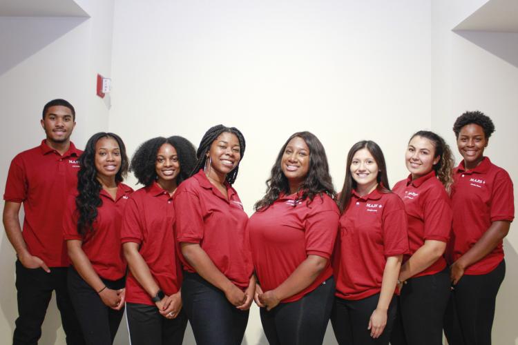 Group of eight students standing in front of white wall with red shirts