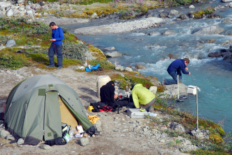 A tent pitched by a clear river with people attending to scientific equipment