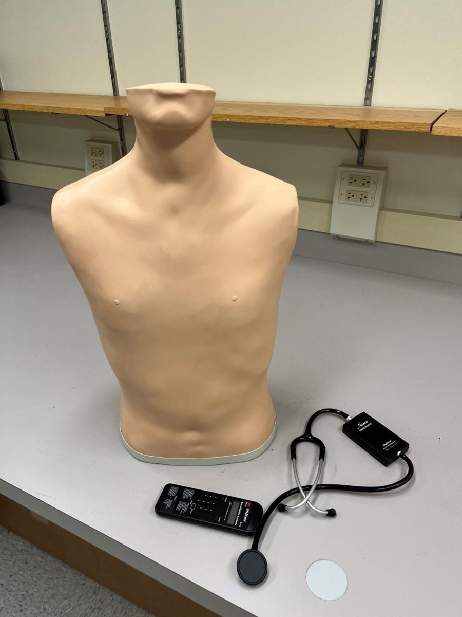 =A manikin torso sitting on a desk with a remote and stethoscope. 
