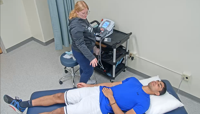 =A student is standing with healthcare equipment beside a volunteer patient on an exam table .