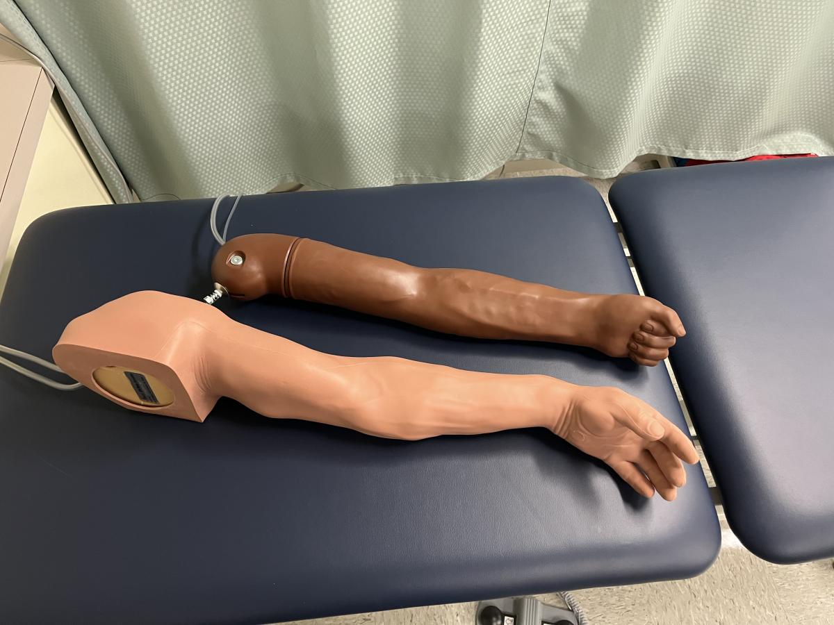 =Two venipuncture trainer arms rest on a blue treatment table with a hospital curtain in the background.
