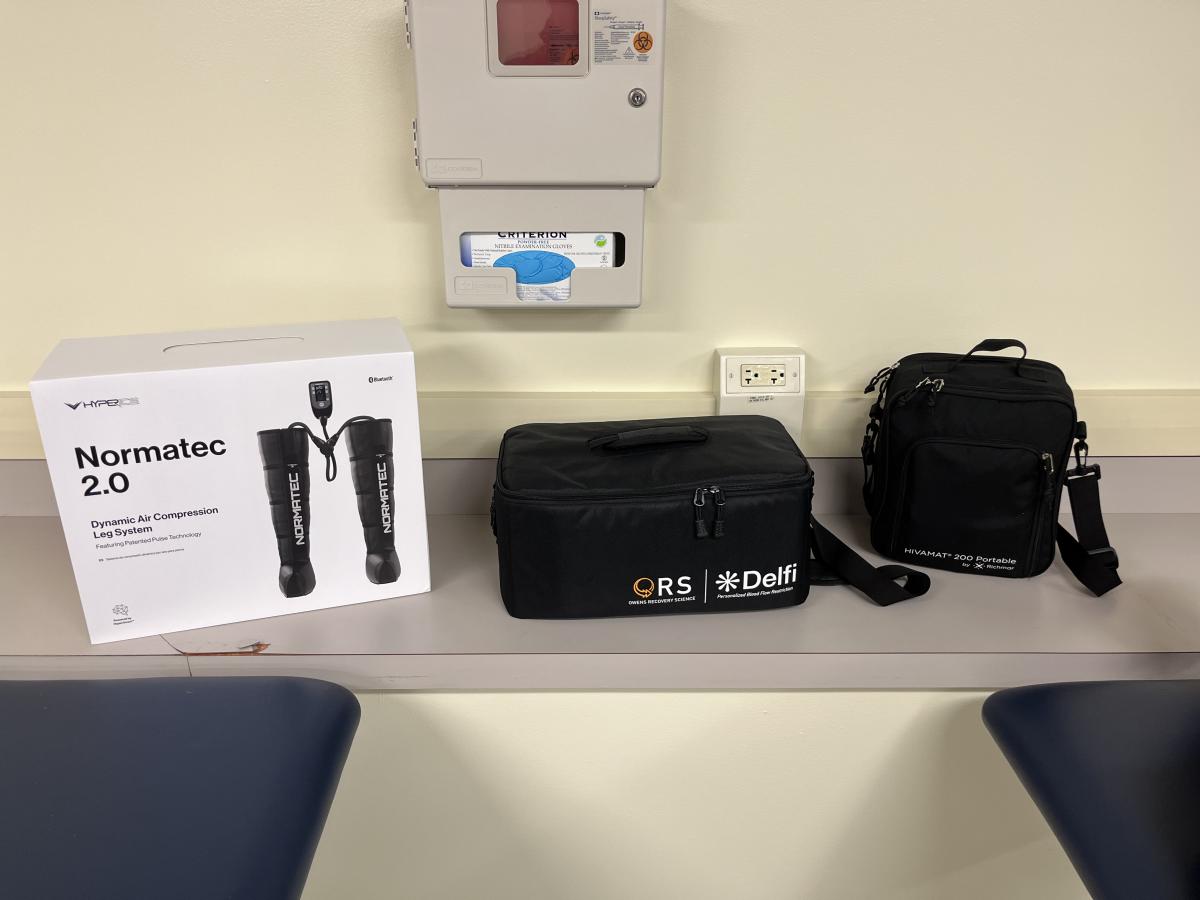 =Pictured on a counter is medical equipment used for athletic training patients. A box of Normatec 2.0 leg compressors, a black bag with ORS Delfi printed on it, and another black equipment bag. The wall has a box for sharps disposal and rubber medical gloves.